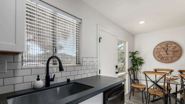 Una cocina moderna con encimera negra, salpicadero blanco de azulejos y grifo negro. Hay una zona de comedor con sillas de madera y un reloj de madera redondo en la pared.