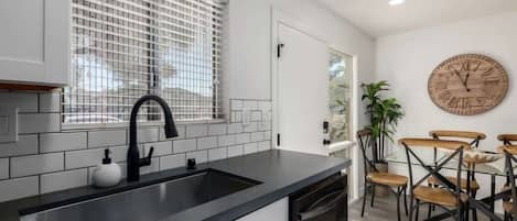Una cocina moderna con encimera negra, salpicadero blanco de azulejos y grifo negro. Hay una zona de comedor con sillas de madera y un reloj de madera redondo en la pared.