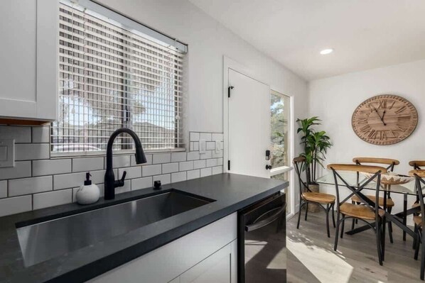 A modern kitchen with a black countertop, white tiled backsplash, and a black faucet. There is a dining area with wooden chairs and a round wooden clock on the wall. - Luxury Camelback House | 4 Heated Pools + Walkable (Scottsdale)