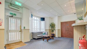 A lobby with a fire exit sign, a sofa, a coffee table, a potted plant, and a fire extinguisher.