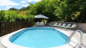 Una piscina rectangular con una escalera metálica, rodeada por un muro de piedra y una valla de madera. Se observan sillones de descanso y una pequeña mesa con sombrilla al fondo.