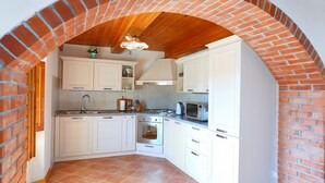 Una cocina con armarios blancos, un arco de ladrillo, techo de madera y suelo de baldosas.