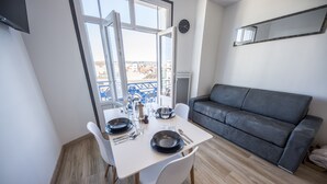 Uma área de refeições com uma mesa posta para dois, um sofá cinzento e uma janela grande com vista para edifícios.