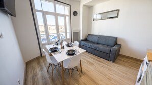 Uma sala de jantar com uma mesa posta para dois, um sofá cinza e uma janela grande com vista para edifícios.