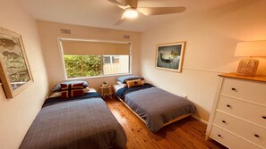 A bedroom with two single beds, a ceiling fan, a window with a view of greenery, a small bedside table, and a dresser with drawers.