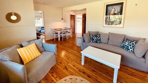 Uma sala de estar com um sofá cinzento, um travesseiro listrado, uma mesa de centro branca e uma área de jantar com uma mesa e cadeiras.