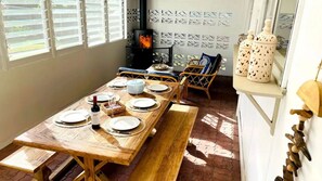 A dining area with a long wooden table set for a meal, a fireplace, wicker chairs, and decorative items on a shelf. - Bombora – Across From The Beach (Callala Beach)