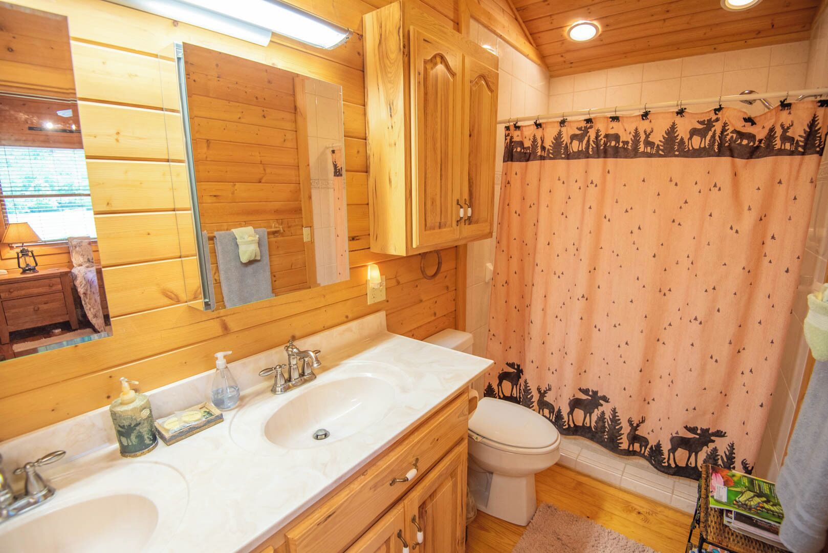 A wooden cabin bathroom with two sinks, a toilet, and a shower curtain with a forest and moose pattern.