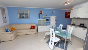 A modern living room with a beige sofa, a glass dining table, and white chairs. There is a kitchen area with white cabinets and a countertop. - Apartment Nautica (Novigrad)