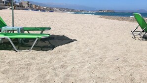 Een strand met groene parasols en stoelen, een heldere lucht en een uitzicht op de zee en verre gebouwen.
