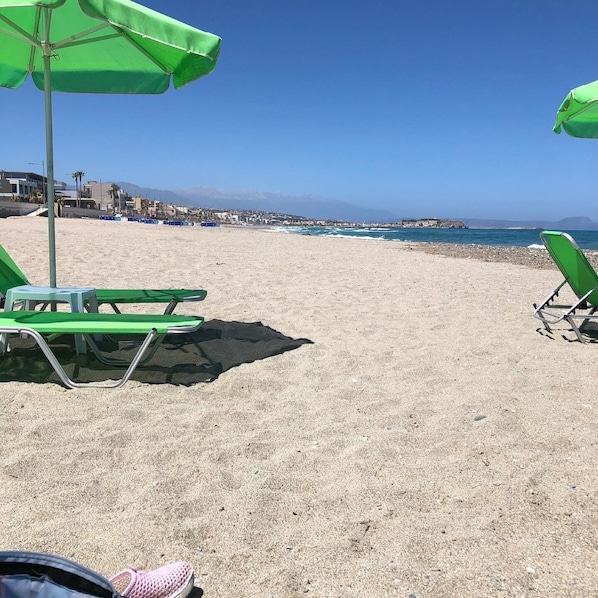 A beach with green umbrellas and chairs, a clear sky, and a view of the sea and distant buildings.