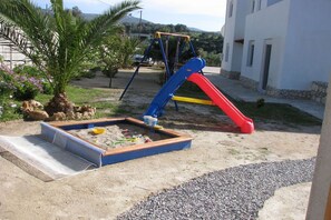 A playground with a slide and swings, surrounded by a garden with palm trees and rocks.