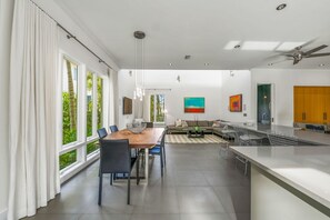 Cozinha moderna com uma ilha grande, área de refeições com mesa e cadeiras de madeira, e sala de estar com sofá e pintura.