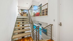 En modern trappa med träpanel och metallräcken, som leder upp till en balkong på andra våningen, där det står en röd stol och hänger en tavla.