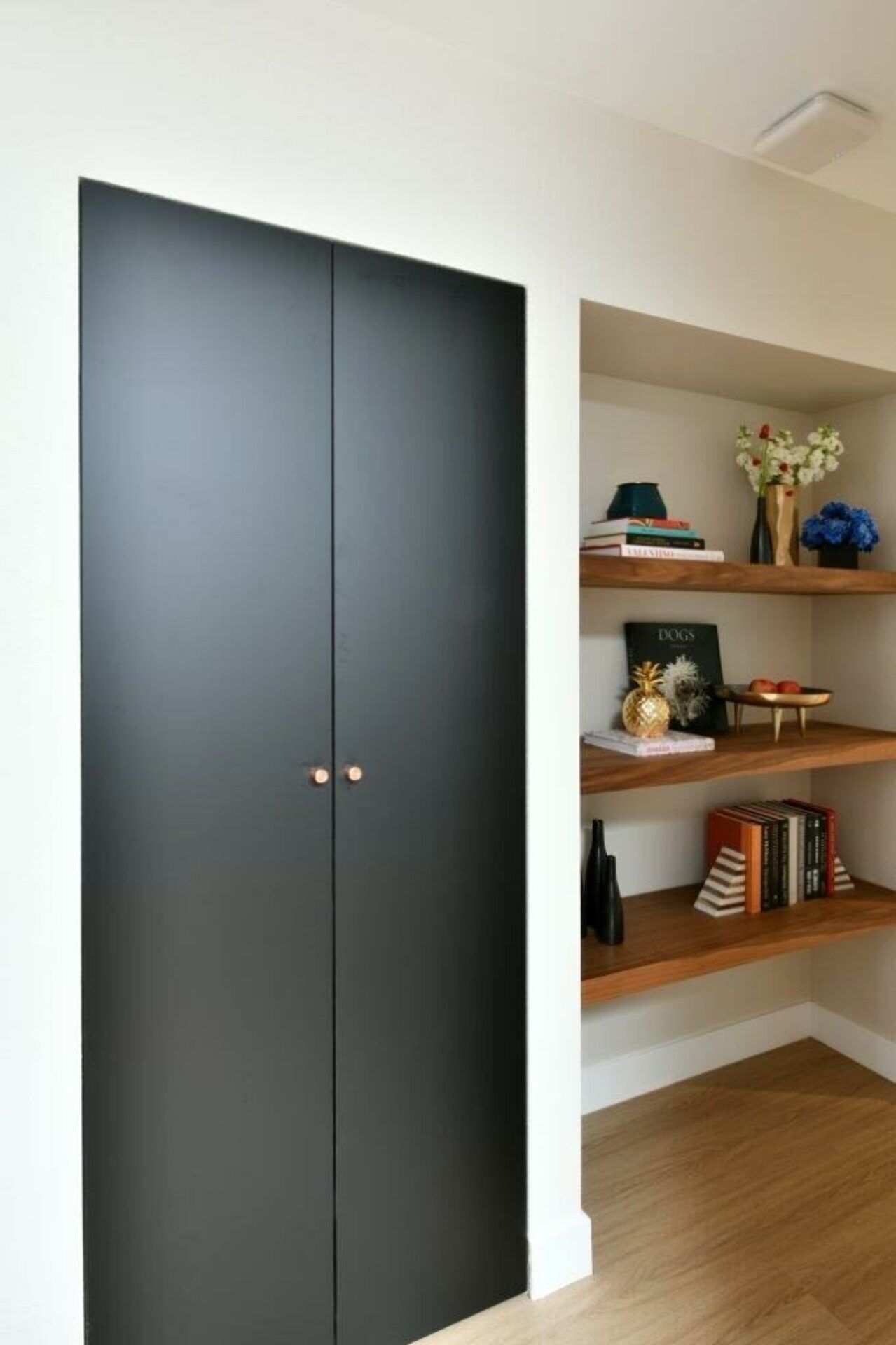 Ein Zimmer mit einem großen schwarzen Kleiderschrank, Holzregalen mit Büchern und Dekoration, und einer weißen Wand.