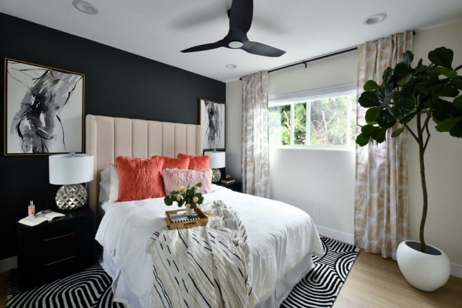 Une chambre à coucher comprenant un lit, une table de chevet, une lampe, un ventilateur de plafond, une fenêtre avec des rideaux et une plante en pot.
