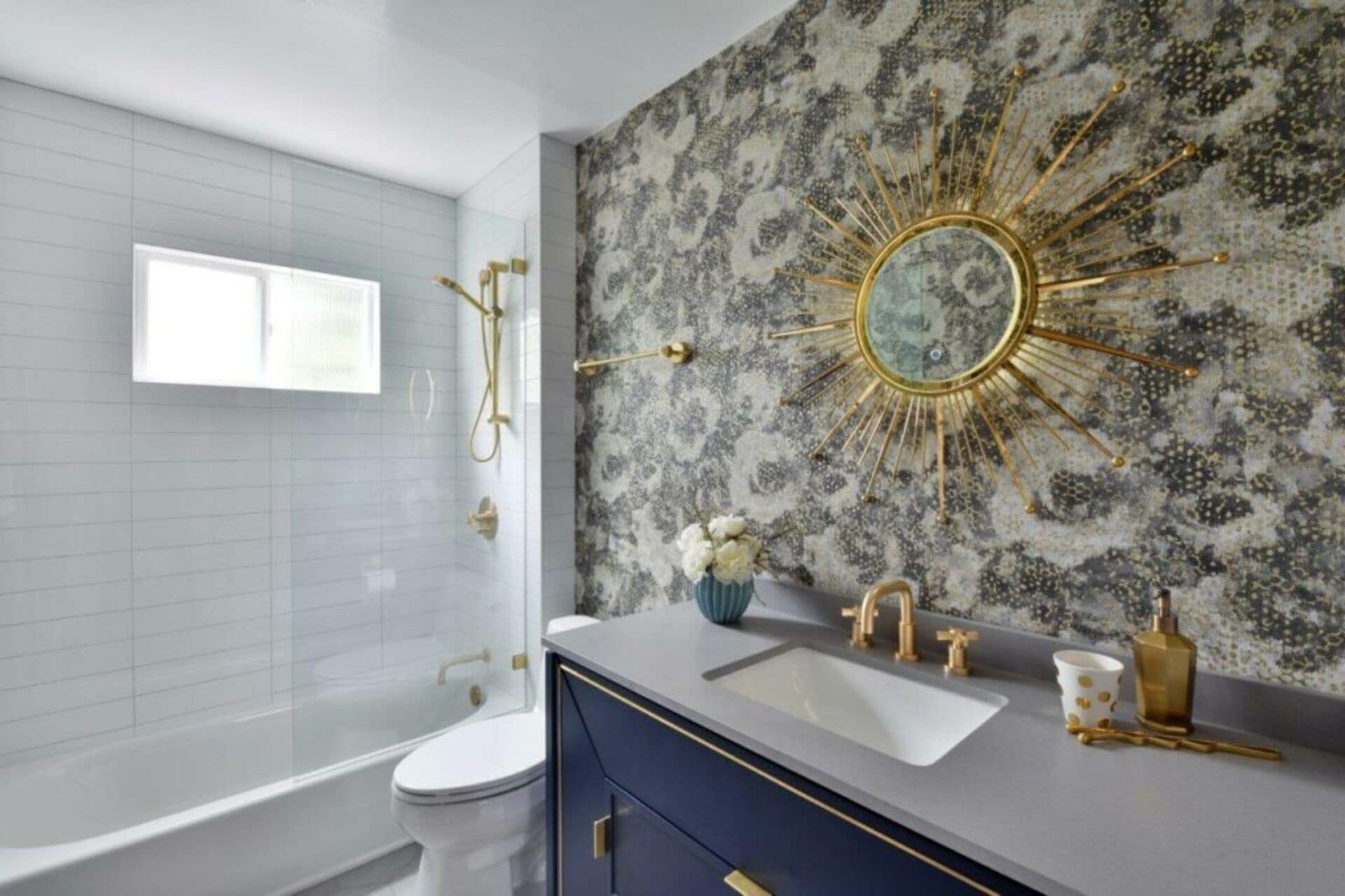 Une salle de bain comprenant une baignoire, des toilettes, un meuble-lavabo et un mur décoratif agrémenté d’un miroir en forme de soleil.