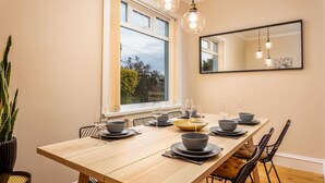 A dining area with a long wooden table set for four, black chairs, and a large window with a view of greenery. - Sunset View - Donnini Apartments (Ayr)