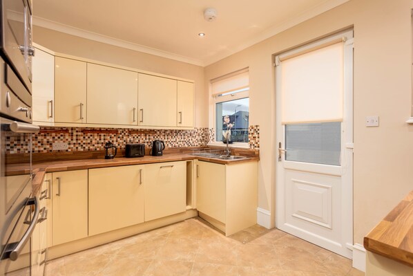 A kitchen with beige cabinets, a tiled backsplash, a wooden countertop, and a window with a view of a building. - Sunset View - Donnini Apartments (Ayr)