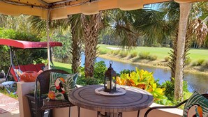 A patio with a round table, chairs, and a red umbrella. A view of a golf course and water is visible through the foliage. - Wildlife Watching! Lagoon & Golf view, beach gear included. Peaceful & Private! (Hilton Head Island)
