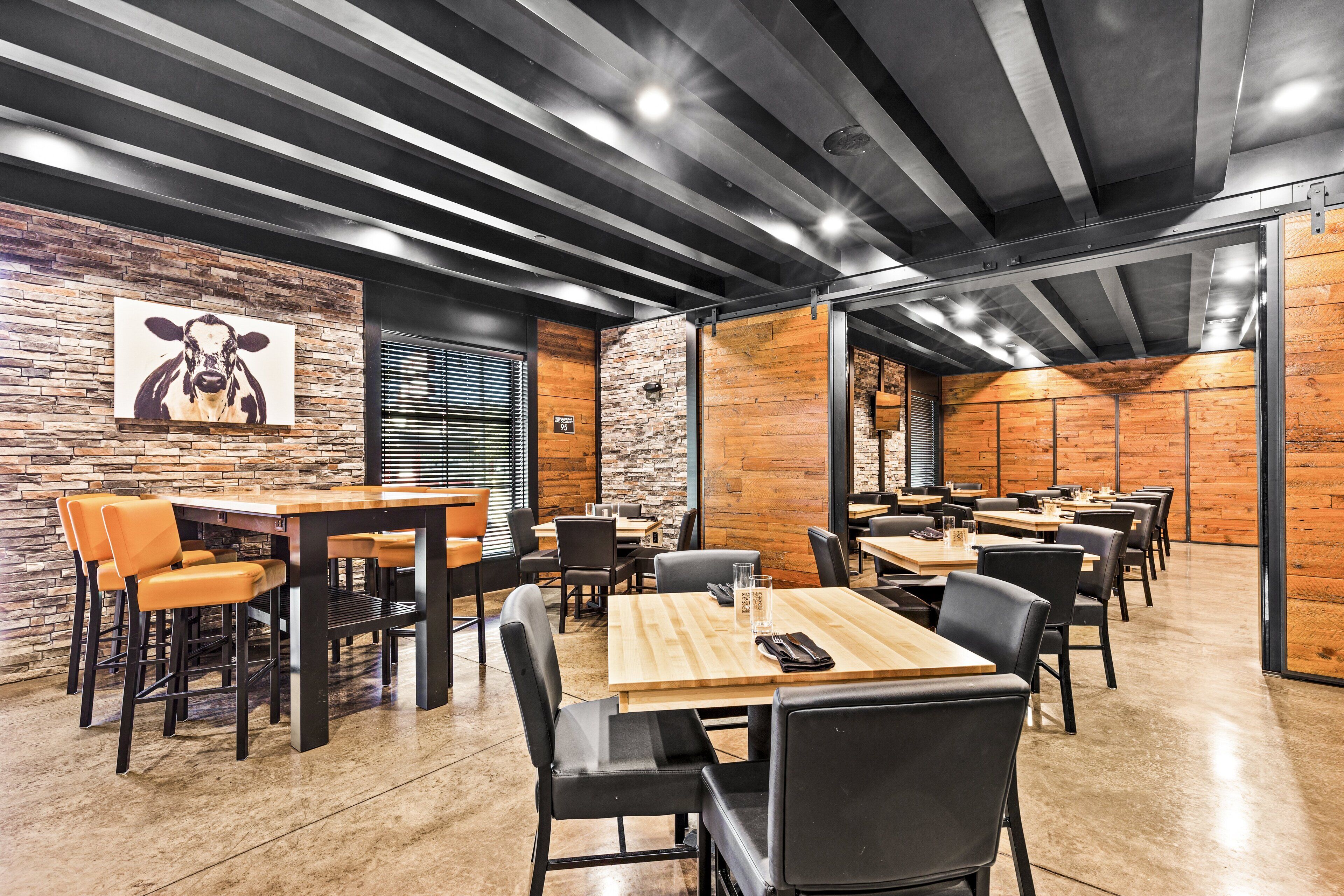 A modern dining area with wooden tables and black chairs, a stone wall with a cow painting, and a brick wall with a wooden panel.