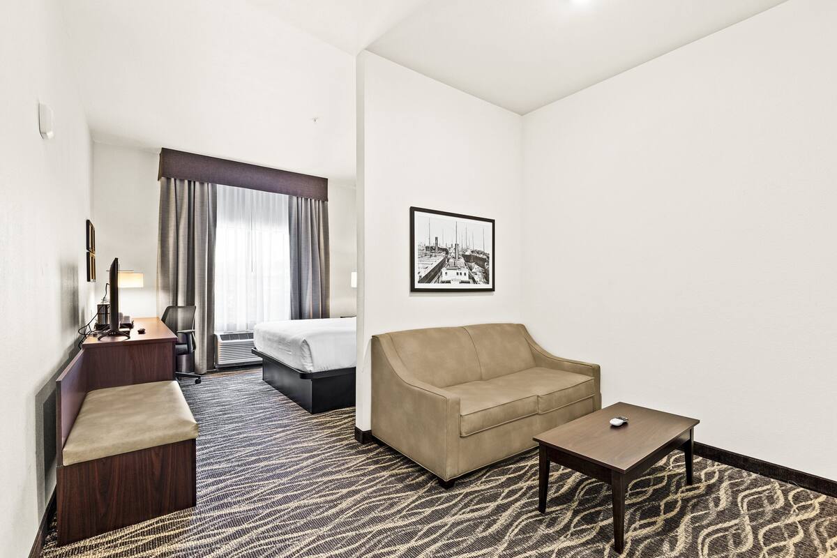 A hotel room with a bed, a sofa, a desk with a lamp, a small table, and a framed picture on the wall.