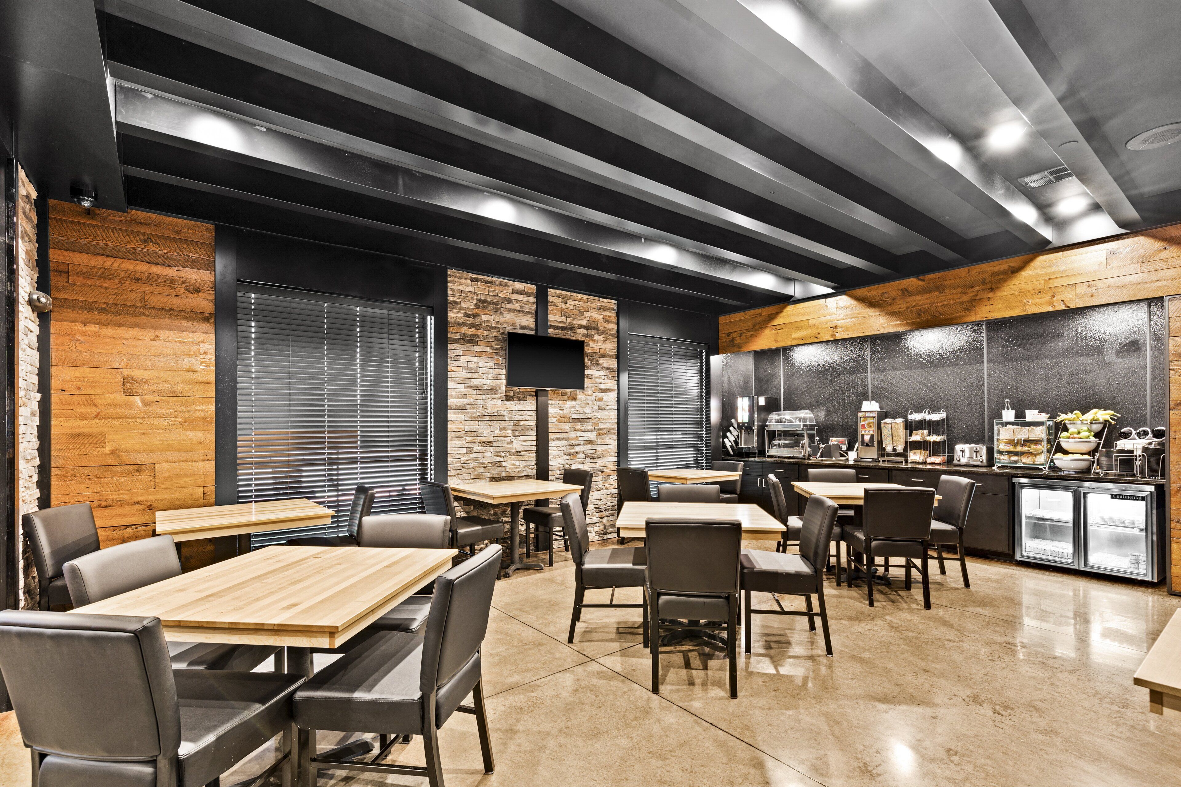 A modern restaurant with wooden tables and black chairs, a flat-screen TV, and a counter with a coffee machine and food display.