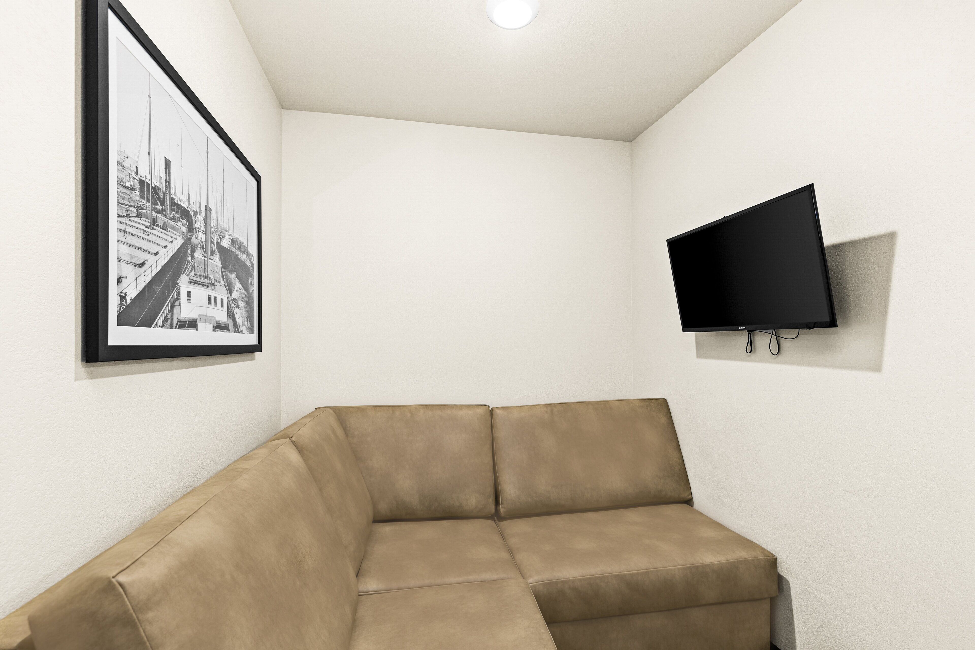 A corner room with a beige sectional sofa, a framed black and white photo on the wall, and a mounted flat-screen TV.