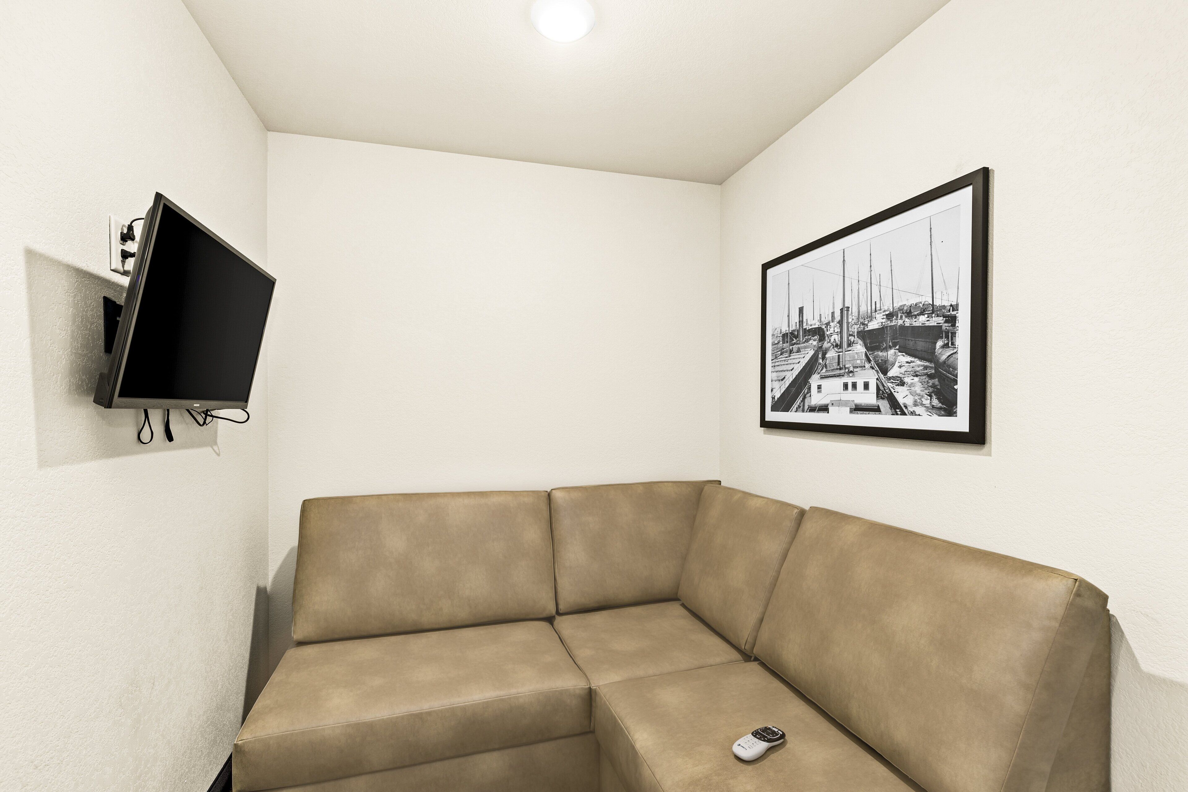 A corner room with a beige sofa, a flat-screen TV mounted on the wall, and a framed black and white photo.