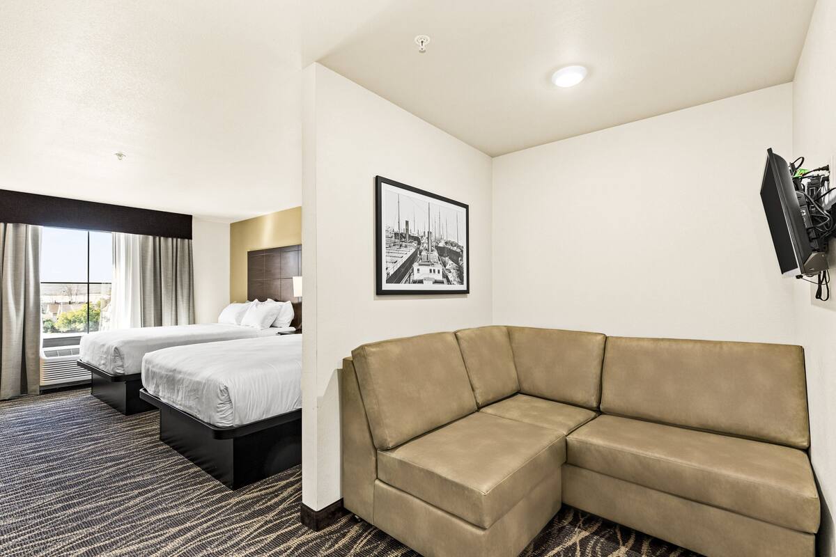 A hotel room with two beds, a sofa, a framed picture on the wall, and a TV mounted on the wall.