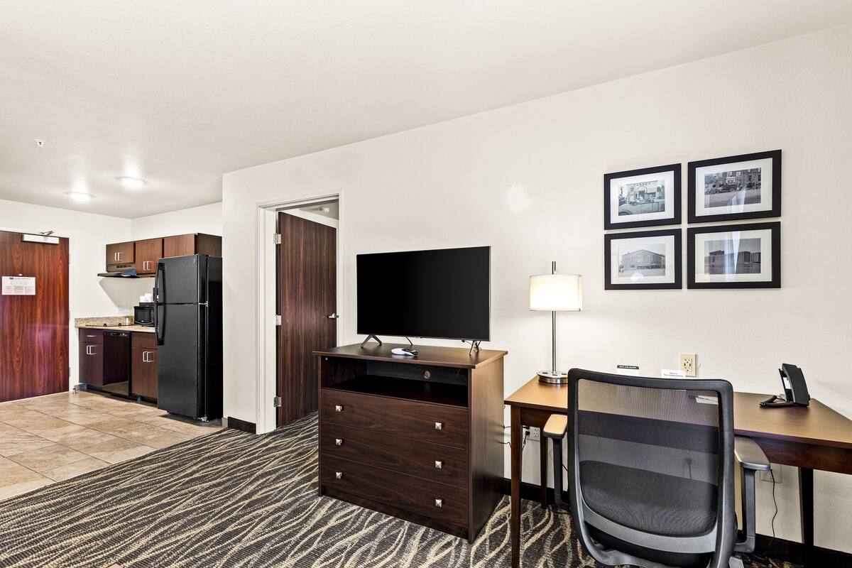 A hotel room with a kitchenette, a desk with a chair, a flat-screen TV, and framed pictures on the wall.