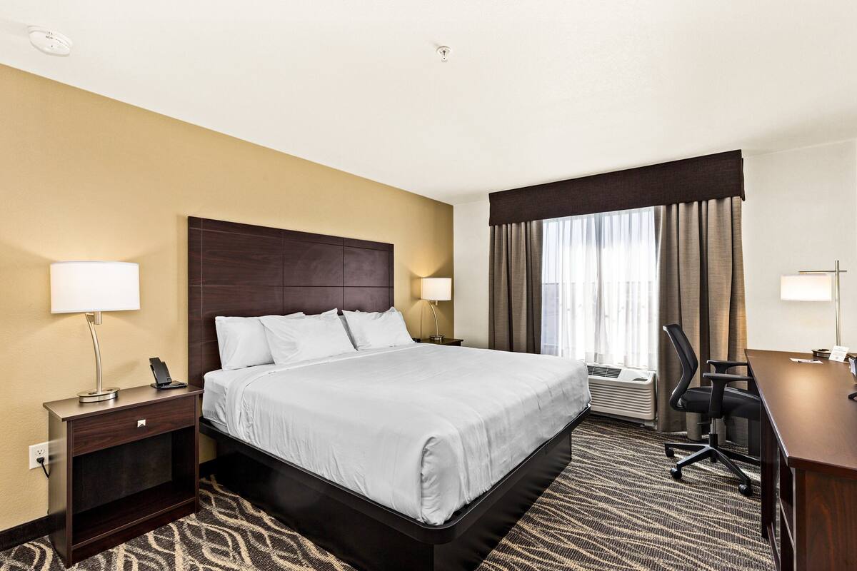 A hotel room with a large bed, bedside tables with lamps, a desk with a chair, and a window with curtains.