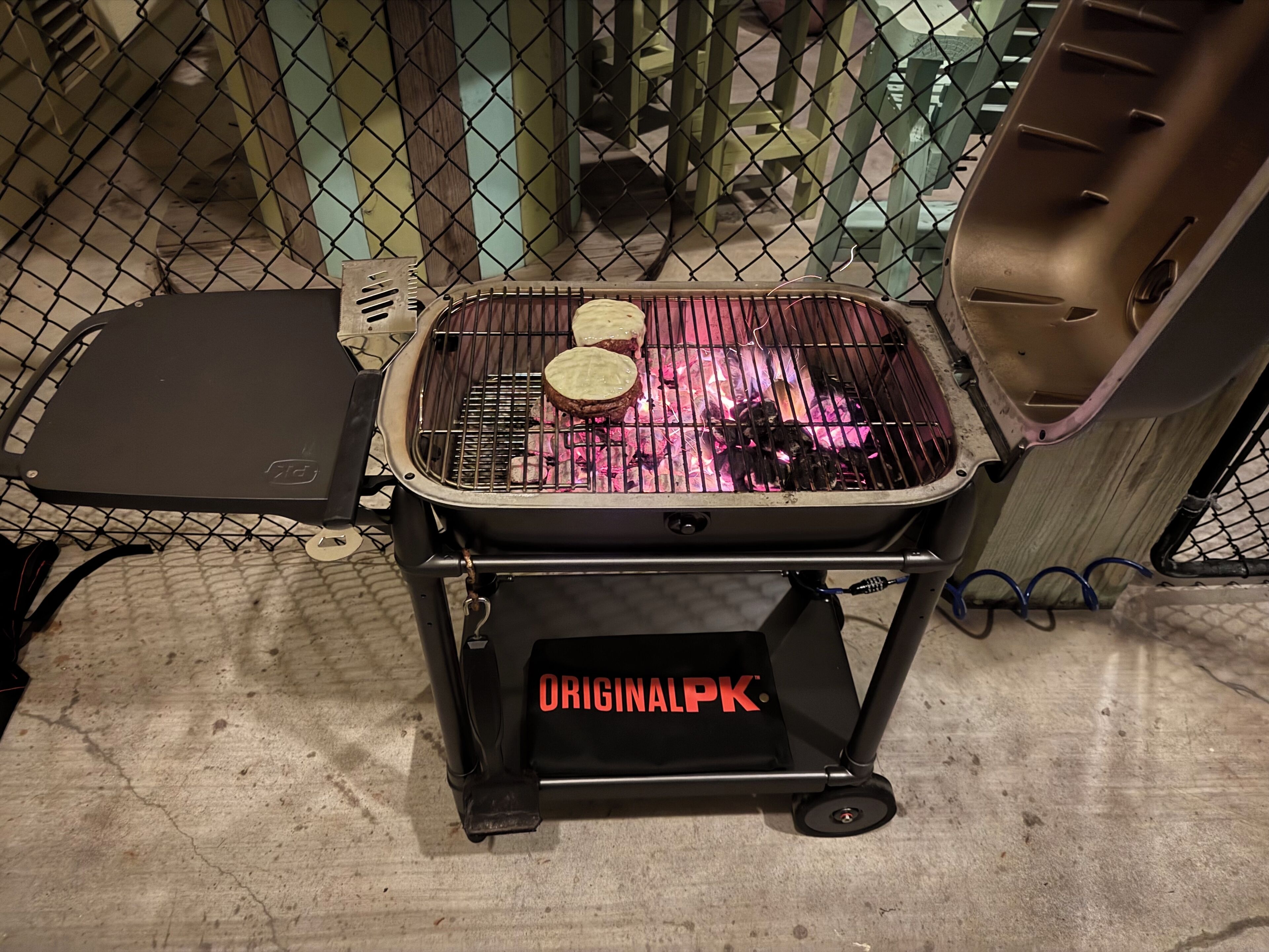 Una parrilla portátil con una rejilla metálica y un asa negra, ubicada sobre un piso de concreto.