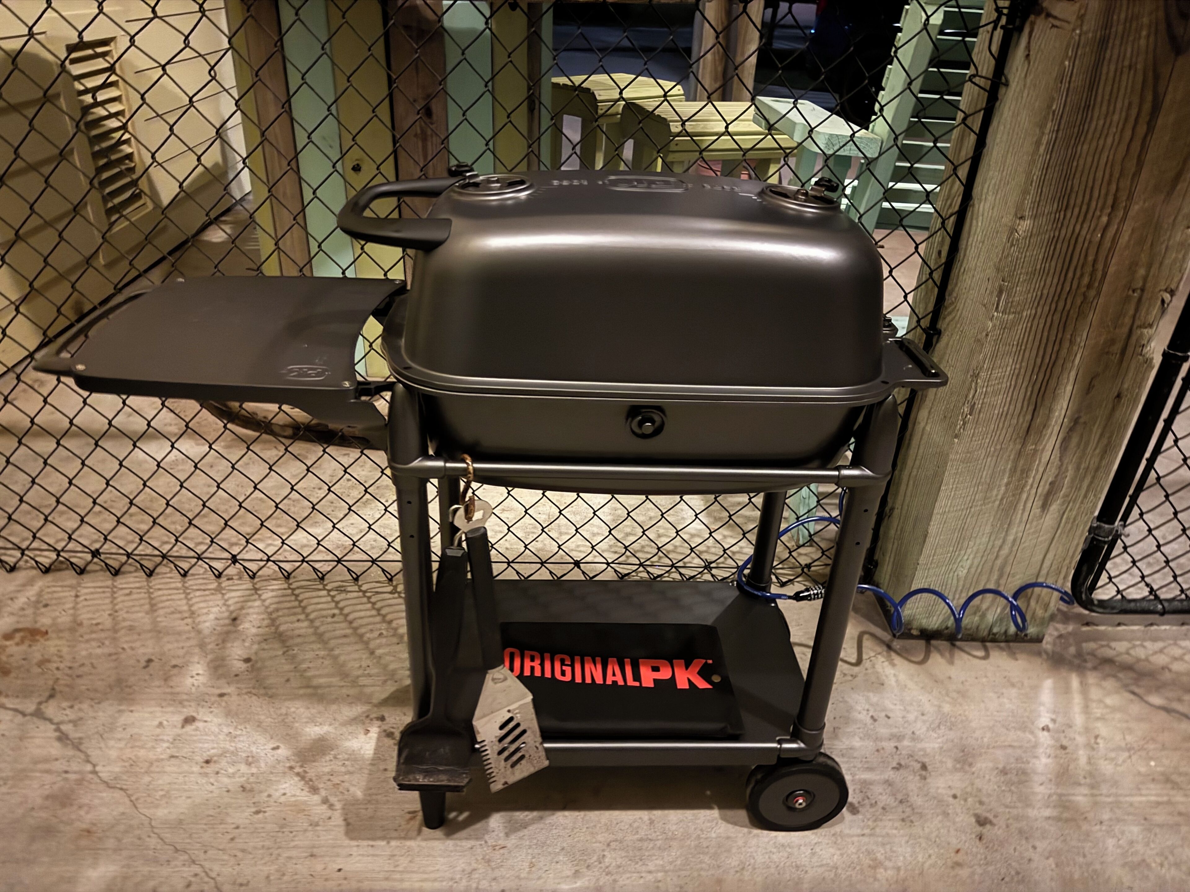 Un barbecue portatif, monté sur un chariot à roulettes, avec une clôture en mailles métalliques en arrière-plan.