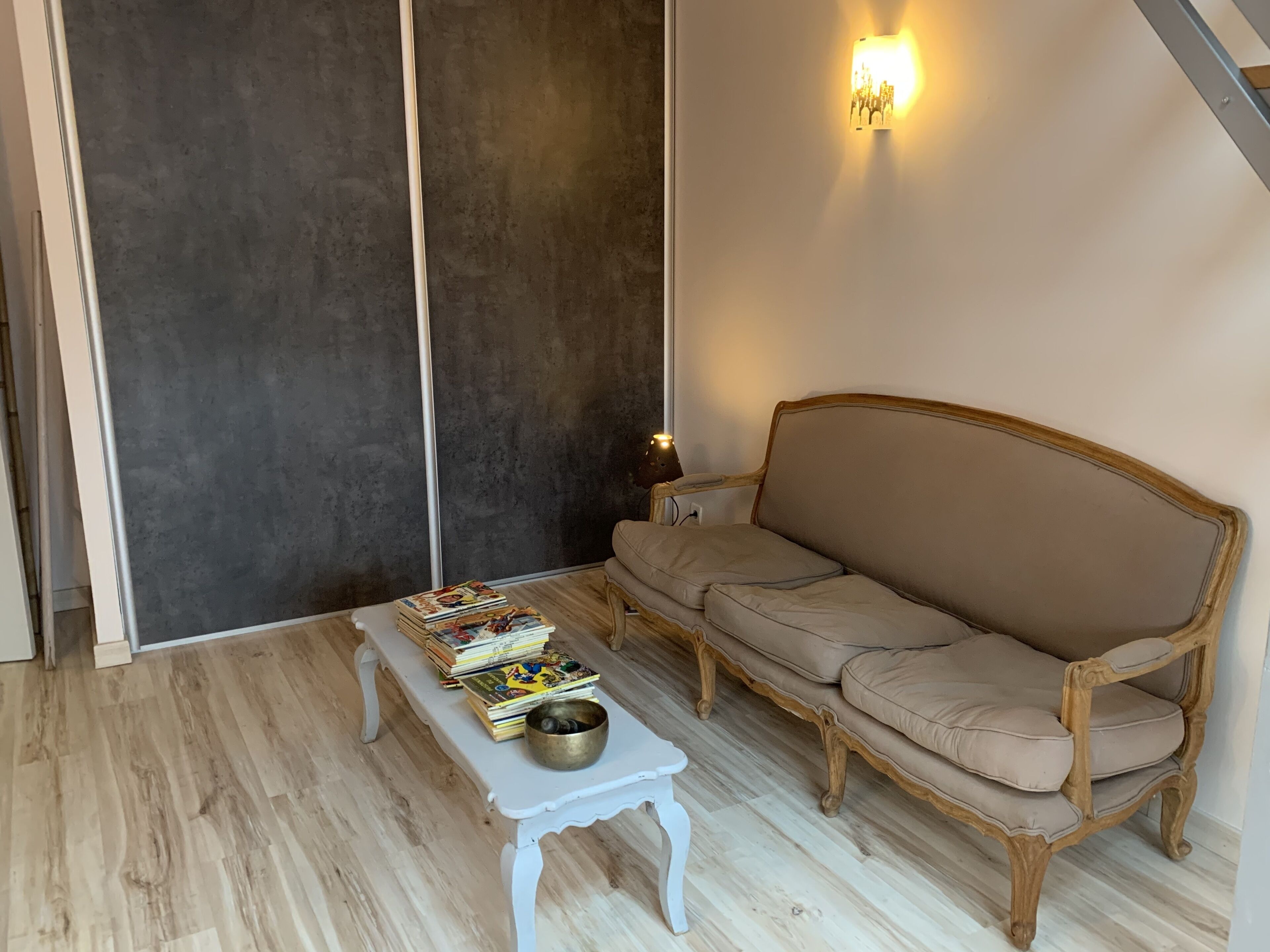 Une pièce comprenant un canapé, une petite table avec des livres et un bol, ainsi qu’un luminaire fixé au mur.