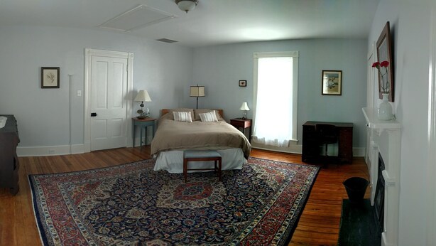 A bedroom with a large bed, a wooden floor, a patterned rug, a dresser, a nightstand with lamps, and a window with curtains. - Welcome Hall 5 bedroom 1880’s home on 55 acres (Paris)