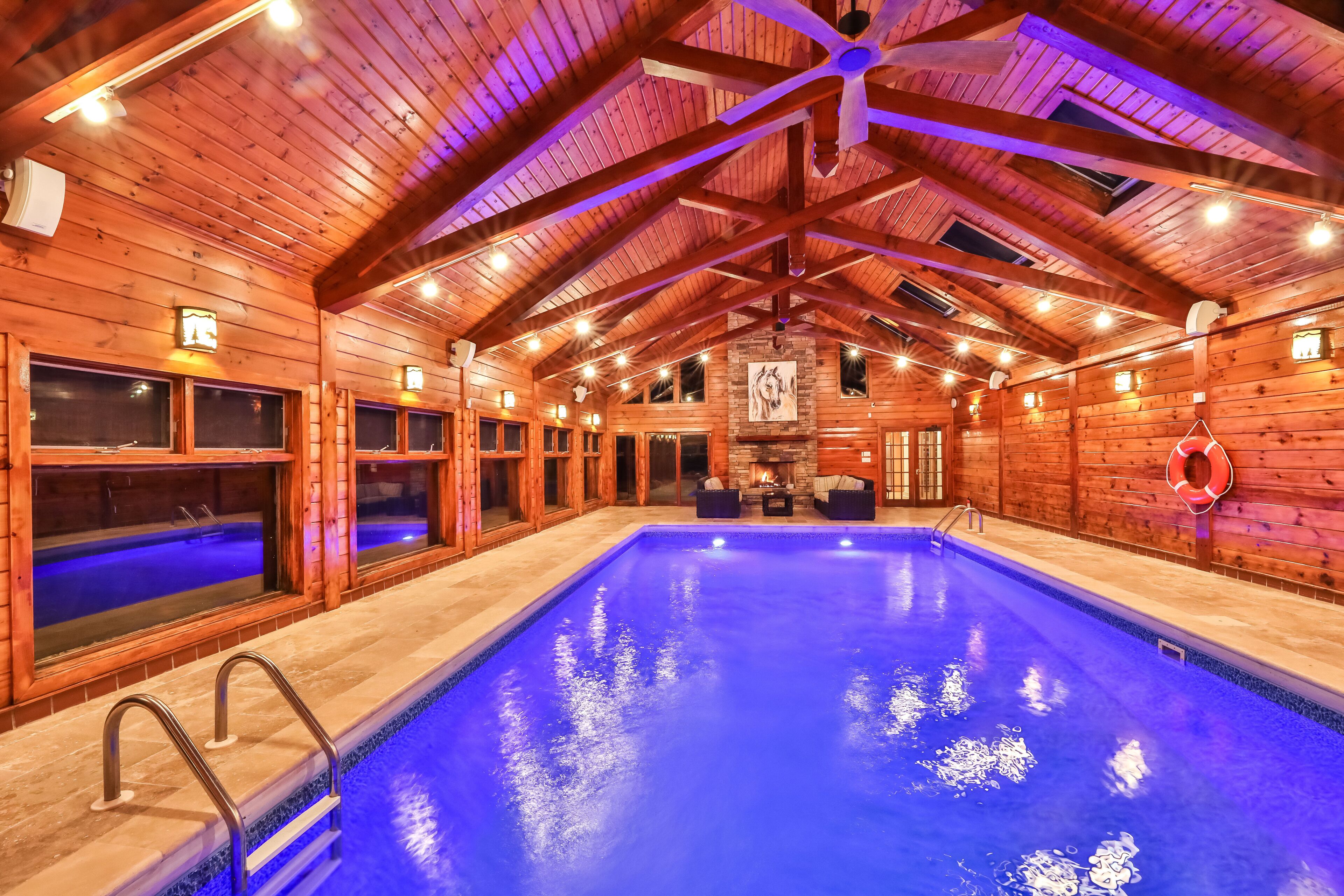 A large indoor swimming pool with a wooden ceiling and walls, a stone fireplace, and a seating area with two chairs.