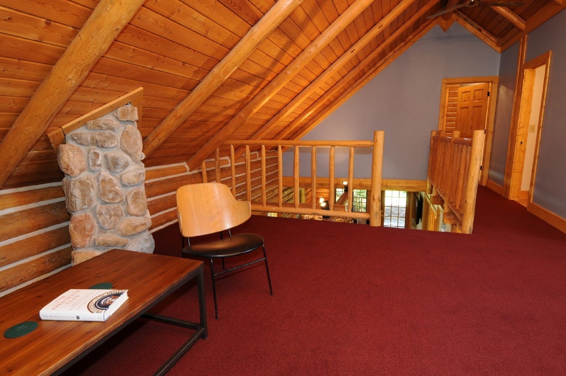 Une pièce mansardée chaleureuse avec un plafond en bois, un mur en pierre, une moquette rouge, une chaise en bois et une table en bois sur laquelle repose un livre.