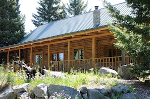 Cozy cabin with a stone chimney and wooden porch, set amidst a natural landscape with flowering plants.