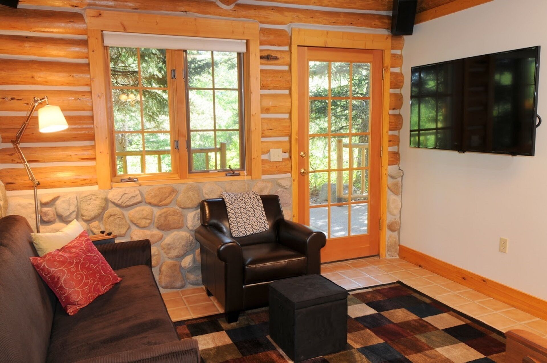 A log cabin interior with a stone wall, a leather chair, a sofa, a lamp, and a television.