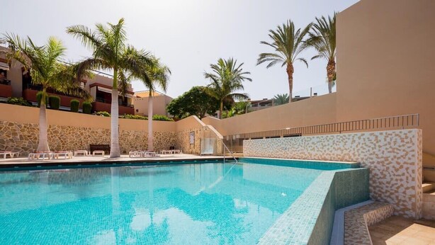 A swimming pool with tiled edges, surrounded by palm trees and a stone wall. - GO2TENERIFE Apart'Tera Solarium, Terraces, Swimming pools & Parking (Adeje)
