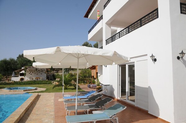 A pool area with lounge chairs and umbrellas, a clear sky, and a white building with balconies. - New - Villa Cornelia with a pool. 2-room apartment. View of the sea above the olive groves (Rethymno - Asteri)