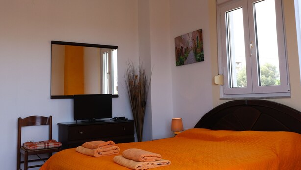 Quarto com cama, televisão, cadeira, penteadeira, janela e um quadro na parede.
