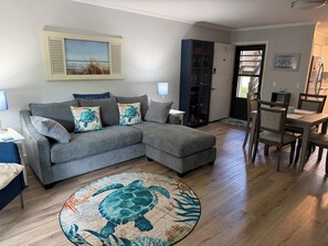Een woonkamer met een grijze modulaire bank, een rond tapijt met een schildpaddes ontwerp, een eethoek met een houten tafel en stoelen, en een keuken zichtbaar door een open deur.