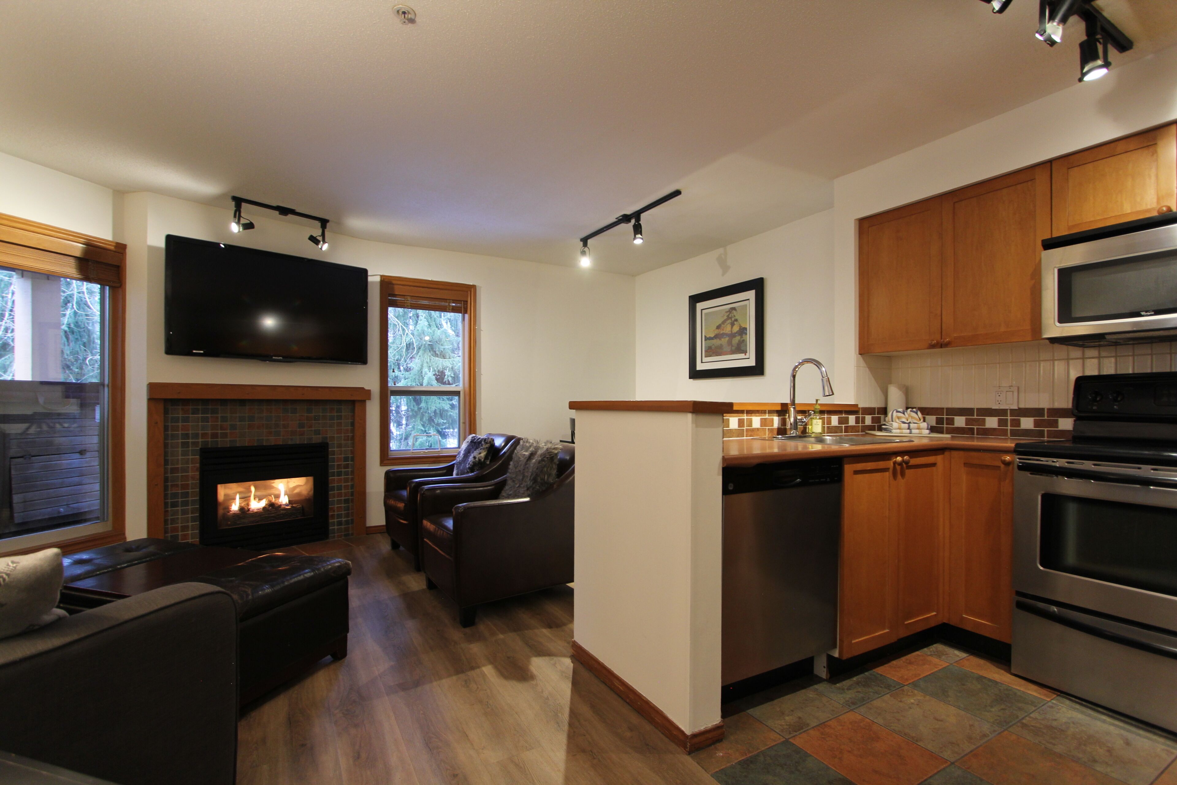 A modern kitchen with a fireplace, wooden cabinets, stainless steel appliances, and a flat-screen TV mounted above the fireplace.