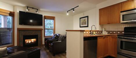A modern kitchen with a fireplace, wooden cabinets, stainless steel appliances, and a flat-screen TV mounted above the fireplace.