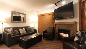 Ein Wohnzimmer mit Sofa, Sessel, Hocker, Kamin und einem an der Wand montierten Flachbildfernseher.