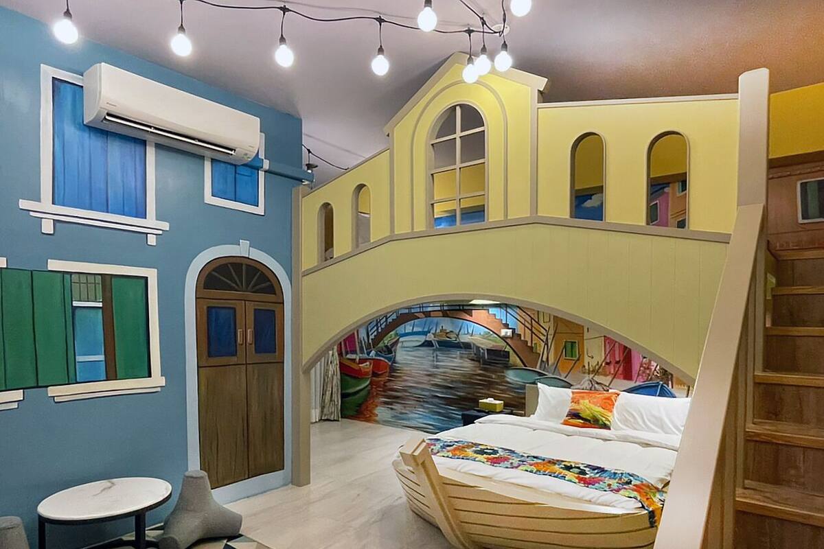 A room with a bed, a small table, and a wall with a mural of a bridge and boats.
