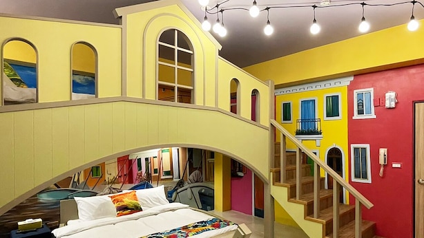 Una habitación con una cama bajo una estructura similar a un puente, una escalera y vistas a un edificio colorido en el exterior.