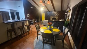 Comedor con una mesa redonda, sillas y individuales amarillos. Zona de bar con taburetes y mostrador. El cuarto tiene pisos de madera y una lámpara decorativa en el techo.
